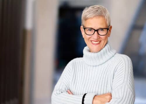 headshot of woman with white cropped hair, black glasses, white knit sweater