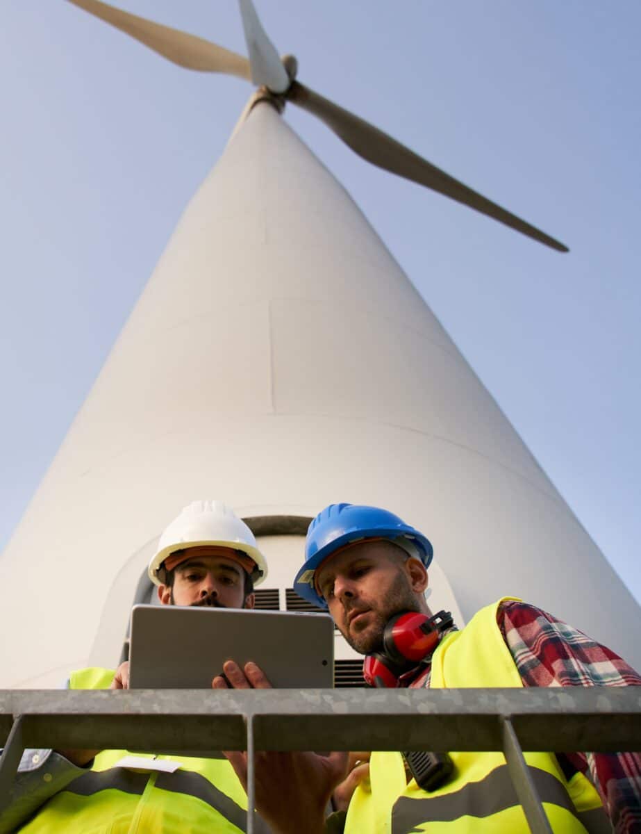 Two helmeted engineers using and looking at tablet. Professional technical workers at wind power plant. People and windmill low angle. Clean renewable energy concept, sustainable future.