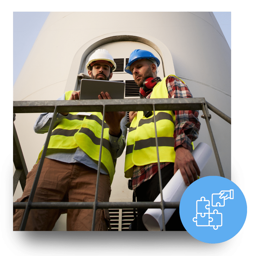 Two engineers in reflective vests and hard hats review plans on a tablet in front of a wind turbine.