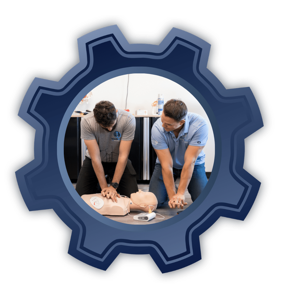 Two people perform chest compressions on a CPR manikin during a training session in a clinical room.