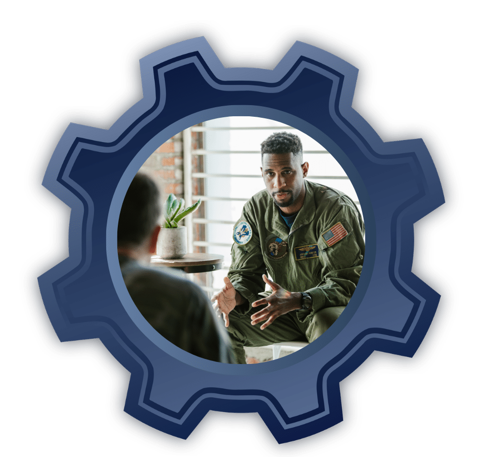 Two servicemen in olive flight suits talk in a casual indoor setting, one gesturing with hands as he explains something