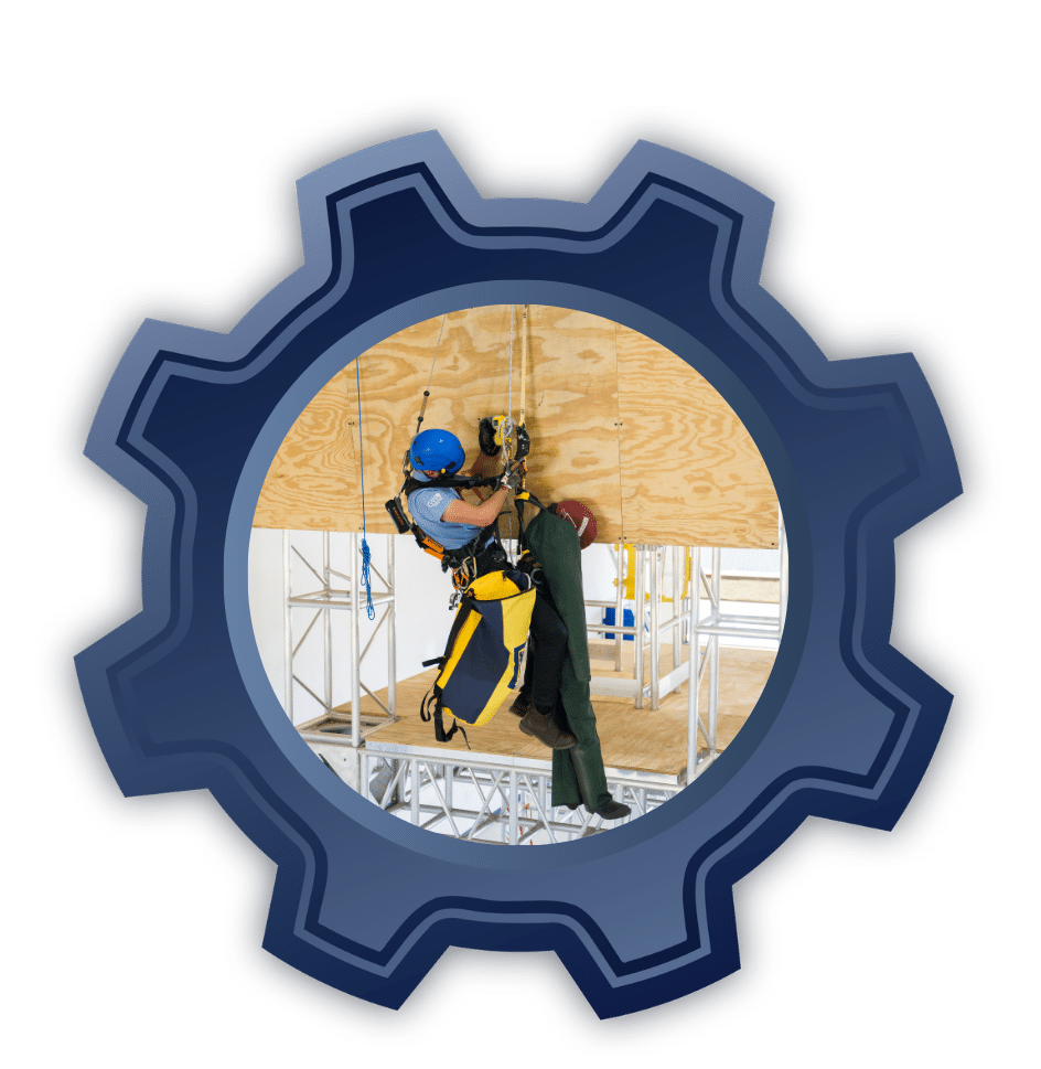 Worker in blue helmet and safety vest practicing rope access on a wooden training wall inside a scaffold frame.